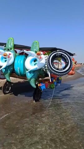 A cart loaded with inflatable pool toys, including several with alligator designs and various swim rings, is situated on a sandy beach at the edge of the water. The cart has two wheels and a canopy. The sea is calm and clear, with gentle waves lapping at the shore. In the background, a few people are visible in the water and on the beach, under a clear blue sky.