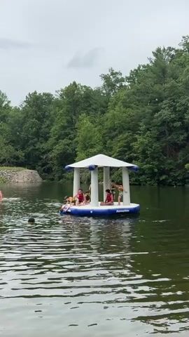 This media file captures a scene on a lake with a large, inflatable water structure in the center. The structure, a white gazebo-like platform on a blue inflatable base, is occupied by several young people, some of whom are wearing life vests. One person is sitting, another is standing, and others appear to be reclining. The inflatable platform has the word "AquaSlide" visible on its side.

Several other individuals are in the water around the inflatable. Two people are seen swimming or floating in the foreground, and another person is partially visible on the left side of the frame, seemingly engaged in an activity on the water. The lake is surrounded by lush, green trees, indicating a natural, outdoor setting. In the background, a rocky outcrop or embankment can be seen on the left. The sky is overcast with clouds, suggesting a diffused daylight and possibly cooler weather.

The overall impression is one of recreational activity on a lake, likely a summer day at a park or a resort. The presence of young people and the inflatable structure point to a fun, leisure-oriented event. The subdued lighting from the cloudy sky creates a serene, yet active, atmosphere. There is no specific indication of the time of day beyond daylight hours. The location is identified as Washington Township, United States of America.