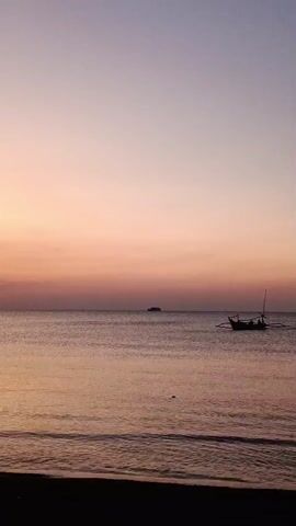 The video shows a wide shot of the ocean at sunset. The sky is a gradient of purple and pink, with a few clouds. The water is calm and reflects the colors of the sky. In the distance, there is a small boat with two people in it. There is also a larger ship further out at sea. The foreground shows a dark beach with gentle waves lapping at the shore.
