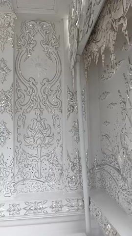 A video shows a close-up view of a white wall adorned with intricate silver and sparkling embellishments. The wall features elaborate ornamental designs, including floral patterns, swirling motifs, and a central figure of a meditating person. The embellishments create a highly decorative and ornate effect, suggesting a temple or a place of worship. The camera pans slowly across the wall, highlighting the detailed craftsmanship and the shimmering quality of the decorations.