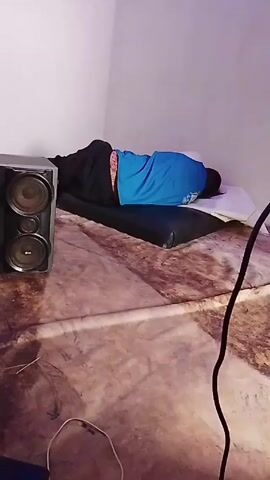 The video shows a person lying face down on a mattress on the floor, with a large speaker next to them. The person is wearing a blue shirt and black pants. The mattress appears to be on top of a larger, patterned sheet or blanket.  A cable is visible in the bottom right corner of the frame. The room has plain white walls.