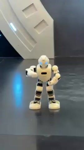 A white, humanoid robot with blue LED eyes stands facing forward.  It has black accents on its limbs and torso.  The robot's arms move from a flexed position to extended, then back again.  The robot is on a gray, shiny surface.  Behind the robot is a curved, white architectural element.  The background is black.  There is a bright blue reflection on the floor in front of the robot.  Music can be heard.