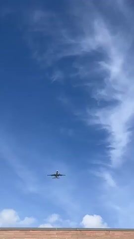 A low-angle shot captures a white airplane ascending into a bright blue sky with wispy white clouds. The airplane is centered in the frame, its belly and landing gear visible as it flies away from the camera. The bottom of the frame shows the top edge of a light-colored structure, possibly a building or rooftop, with horizontal lines. The overall impression is one of clear weather and a plane in flight.