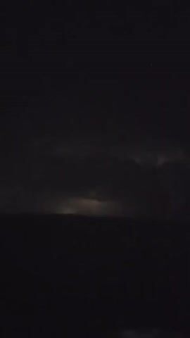 A time lapse video of a distant thunderstorm displays brief flashes of lightning illuminating the dark, cloudy sky over a low horizon line. The scene is almost entirely dark, with the lightning flashes being the only significant source of light, momentarily revealing faint cloud formations. The overall impression is one of a powerful natural event occurring at a distance.