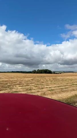 A red vehicle's hood is visible in the foreground of the video, partially obscuring a wide shot of a harvested field. The field displays neat rows of cut crops, appearing golden brown and dry. In the distance, a line of trees creates a dark green contrast against the horizon. The sky above is a vibrant blue, dotted with fluffy white clouds that transition to darker, more ominous gray clouds towards the right side of the frame. As the video progresses, hay bales become visible scattered across the field, and several wind turbines appear on the distant horizon. The camera pans slightly, offering a view of a dirt path on the right side of the field.