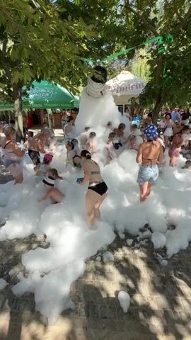 A group of people, including adults and children, are playing in a large pile of white foam. The foam appears to be generated by a machine that is visible at the top of the pile, with foam pouring out of a large nozzle. The scene takes place outdoors, under trees, with some tents and buildings visible in the background. People are dressed in swimwear and appear to be enjoying themselves. Some individuals are submerged in the foam, while others are standing or sitting in it.