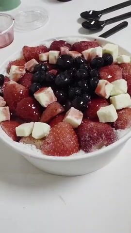 A bowl of shaved ice topped with strawberries, blueberries, and chunks of white cheese. The shaved ice is fine and powdery. The strawberries are sliced and arranged around the bowl, with some whole blueberries scattered on top. Small pieces of white cheese are interspersed with the fruit. A drizzle of red syrup covers some of the strawberries and blueberries. The bowl is white and appears to be paper or styrofoam. In the background, there are two black spoons and a small clear container with a pink lid. The scene is set on a white surface, possibly a table.
