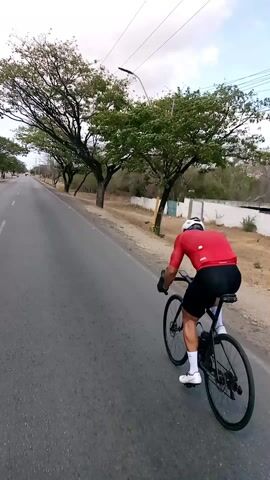 A cyclist is seen from behind, riding down a paved road lined with trees. The cyclist is wearing a red short-sleeved shirt, black cycling shorts, white socks, and white cycling shoes, and a white helmet. The bicycle is black. The road has some markings on the side, and there are power lines and poles visible above. The environment appears somewhat rural, with trees and vegetation on the side of the road. There's a white wall in the background on the right side. The sky is overcast.
