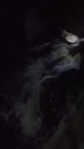 The video shows a dark, outdoor scene with a waterfall. The water is white and foamy as it cascades down. There are rocks visible in and around the water. The lighting is dim, suggesting it is nighttime or very early morning. There are no people or animals visible in the video. The environment appears to be natural and undeveloped.