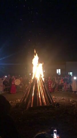 A large bonfire burns in the center of a dark outdoor space. Sparks fly upward from the flames. A crowd of people surrounds the bonfire. Some people in the crowd are wearing traditional clothing, including robes and headdresses. Some individuals are playing drums. The people appear to be observing the bonfire, and some are holding up cameras to record the event. The ground around the bonfire is dirt and rocks. In the background, there is a building with lit windows. The sky is dark and appears to be nighttime.