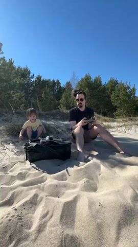Here's a description of the video:

A man and a young child are on a sandy beach on a sunny day. The man is sitting cross-legged, operating a drone with a controller. The child, wearing a yellow shirt and blue shorts, is initially sitting near the man, then stands up and reaches for the drone as it takes off. A black bag sits between them on the sand. There are trees in the background. The sky is clear blue.