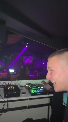 A person in a black T-shirt with a colorful palm tree graphic is seen at a DJ booth, while another person in a black T-shirt with "BALENCIAGA" written on it is dancing in front of them. The DJ booth has various equipment, including a mixing console and laptops, all bathed in purple and green lighting. Disco balls hang from the ceiling, reflecting laser lights, creating a vibrant club atmosphere.
