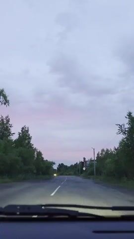 The video is filmed from the perspective of someone driving a car. The camera is looking forward through the windshield, which has wipers at the bottom. The road ahead is asphalt and has a white line down the center. There are trees and bushes on either side of the road. The sky is cloudy and appears to be dusk or dawn, with a pinkish hue on the horizon. In the distance, another car with its headlights on is approaching. There is a traffic sign on the right side of the road and a billboard further down. The lighting suggests it is not full daylight.