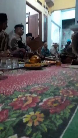 The video shows a gathering of men, likely elders, seated on the floor in a room. They are dressed in traditional attire, including peci hats. In front of them, on a colorful floral carpet, are plates of food and drinks, suggesting a meal or a communal gathering.

The scene appears to be indoors, possibly a home, with a doorway leading to another area visible in the background. The lighting suggests it could be evening or an overcast day. The atmosphere seems calm and respectful, with the individuals engaged in conversation or a shared activity. One person is holding a book or a large piece of paper, which might be related to the gathering's purpose.

The location is identified as Panjalin Kidul, Indonesia, which provides cultural context for the attire and activities. The food items on the plates are not clearly identifiable due to the video's resolution, but they appear to be small portions, possibly snacks or part of a larger meal. There is a visible inscription on a yellow pot in the center of the table, which reads "BUAN", but the full context is unclear. The overall impression is of a traditional gathering, possibly a religious or social event, where community and tradition are being honored.
