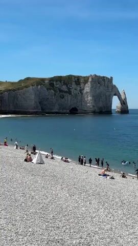 The video pans across a rocky beach under a bright blue sky. In the background, a large cliff formation with a natural archway stands in the blue ocean. Several boats, including a small motorboat and what appear to be kayaks, are visible in the water. On the beach, numerous people are scattered about. Some are sunbathing, others are walking, and a group is gathered near the water's edge. A few individuals are in the ocean, swimming or wading. Towards the right side of the frame, a beach umbrella provides shade for two people. The overall scene depicts a lively beach day with people enjoying the sun and the sea.