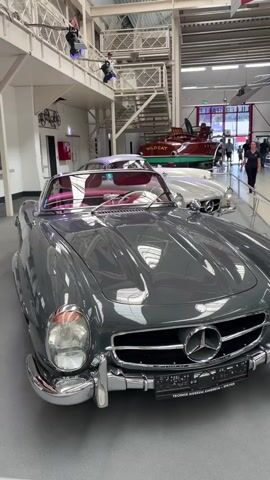 The video pans across a display of vintage cars in what appears to be a museum. In the foreground, a dark gray Mercedes-Benz 300 SL Roadster is prominently featured, its shiny exterior reflecting the overhead lights and structure of the building. Its interior, with plush pink upholstery, is visible. Further back, a lighter colored Mercedes-Benz, possibly another 300 SL, is parked behind it. In the background, a red speed boat with "WILDCAT" written on its side is displayed. The environment is spacious with high ceilings, exposed beams, and walkways or mezzanines above. Several people are visible in the background, casually observing the exhibits. The lighting is bright and artificial, typical of an indoor exhibition space. The camera movement is smooth, following the lines of the cars.