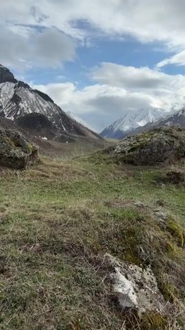 The camera pans across a mountainous landscape.  In the foreground are two large, lichen-covered rocks framing a view into a valley. The valley floor is green with a visible road running through it. Towering snow-capped mountains rise in the background, partially obscured by a sky filled with white, puffy clouds. The overall lighting suggests a cloudy day with patches of sunlight.