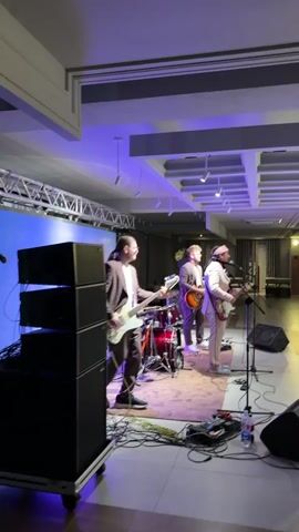 A band is performing on a stage. There are four band members visible. The person on the far left is playing a white bass guitar and wearing a brown suit and white shirt. The person next to them, in the middle, is playing a red drum set. The person to the right of the drummer is wearing a light-colored suit and playing a guitar. The person on the far right is wearing a light-colored outfit, possibly a suit, and a pink bandana, and appears to be singing into a microphone while playing a guitar. There are speakers on the left and right of the stage, and lighting equipment above. A large screen is behind the band, displaying a colorful abstract pattern. The floor is tiled, and there are numerous cables running across the stage.
