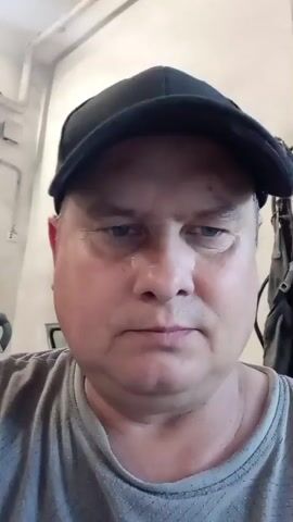 This is a close-up, selfie-style video of a man wearing a black baseball cap and a grey t-shirt. He appears to be indoors, in what could be a workshop or a garage, given the background elements such as metal pipes, a vent on the wall, and general industrial-looking surroundings. The lighting suggests it might be daytime, but the interior setting makes it difficult to be certain. The man's expression is neutral, and he is looking directly at the camera. There are no specific events or activities clearly taking place; the focus is solely on the man's face and upper body. The video's context of Jablonec nad Nisou, Czechia, provides a geographical location but doesn't offer specific visual cues within the frame itself. No text is visible in the media file.