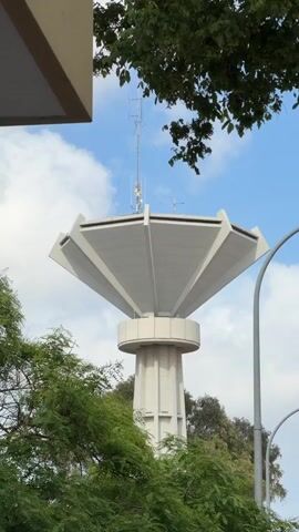 A vertical shot captures a distinctive concrete water tower in El Prat De Llobregat, Spain, against a blue sky with white clouds. The tower features a thick, cylindrical base that supports a wider, multi-faceted top section, resembling a large, inverted umbrella or flower. This top structure is light grey and has prominent angular segments.

The camera initially focuses on the mid-section of the tower, then slowly pans upwards, revealing the very top. Several communication antennas of various sizes and types are mounted on the highest point of the tower, indicating its dual function as a water reservoir and a communication hub. As the camera continues to pan, it moves past the tower's top, revealing more of the sky. A small, dark bird is seen flying from left to right across the frame, silhouetted against the bright clouds. Green foliage from trees is visible in the foreground and partially obscures the lower parts of the tower, as well as a section of a dark grey lamppost or pole on the right. The lighting suggests it is daytime.