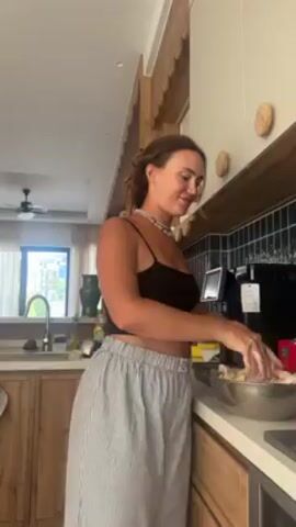 A woman is standing in a kitchen, mixing ingredients in a silver bowl. She is wearing a black tank top and striped wide-leg pants. The kitchen has wooden cabinets, white walls, and a black tiled backsplash. A window in the background shows greenery outside. The woman is looking down at the bowl and talking.