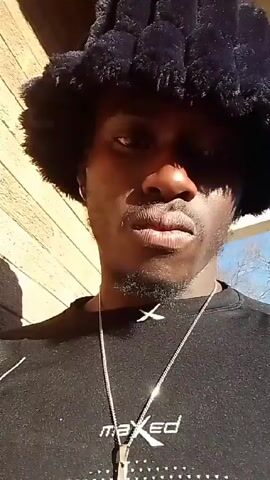 Here is a description of the video:

The video shows a close-up of a person facing the camera. The person has dark skin, a short beard, and wears a black, fuzzy bucket hat. The hat casts a shadow over their forehead.  They are wearing a black long-sleeved shirt with a small white "X" logo near the collar and a silver chain with a cross pendant. The background appears to be a light brick wall, partially in the sun. The person has a serious expression, and the camera is steady. The video also has background music.