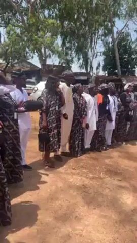 A group of people standing in a line outdoors are addressed by a person in white clothing. The people in the line are wearing a mix of white clothing and patterned clothing that is black and brown. Most of the people in the line are wearing some kind of identification badge. The line of people is standing on a dirt patch under the shade of a group of trees. Some low buildings and a parked car are visible in the background. The person in white motions and points at the line of people.