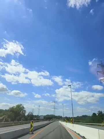 The video shows a point-of-view recording from inside a vehicle driving on a highway. The sky is a bright blue with scattered white clouds. Several tall, lattice-like electricity pylons are visible in the distance. The road has multiple lanes and is bordered by a concrete barrier. There are a few other vehicles on the road. The camera pans slightly upward, capturing more of the sky and the power lines. Some small, dark objects, possibly birds or debris, are visible in the sky. There are also a few orange spherical objects suspended from the power lines, which appear to be markers. The overall environment suggests a sunny day on a major road.
