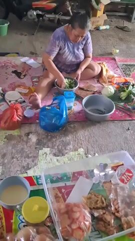 A woman is sitting on a mat on a concrete floor, preparing food. She is wearing a tie-dye shirt and shorts. In front of her is a bowl of chopped green vegetables. To her right are several bunches of green vegetables, a white bowl, and a plastic bag. To her left are a red plastic bag, a small blue container, and a small blue bag. In the foreground, there is a clear plastic container filled with various food items, including sliced meat, nuts, and crackers. In the background, a motorcycle is parked. The scene appears to be outdoors, possibly in a garage or a covered outdoor area.