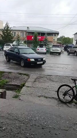 A black Audi sedan parked on a wet asphalt surface. In the background, several other vehicles are visible, including white SUVs, a silver minivan, and a dark-colored sedan. A bicycle is parked on the side of the road. Buildings are visible in the background. The weather appears to be rainy, with wet roads and a cloudy sky.