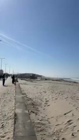 The video shows a sandy beach under a clear, bright blue sky with condensation trails of a plane. A concrete walkway runs along the edge of the beach with a few people walking on it. The sand is marked with footprints and tire tracks. Waves break on the shore in the distance.