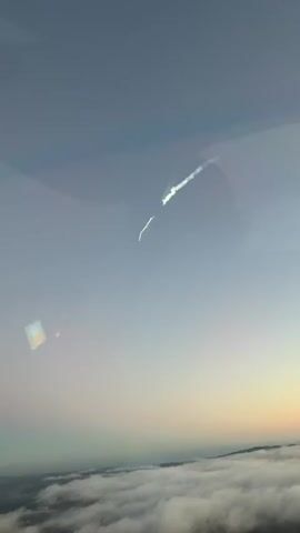 This is a video of a rocket launch, likely from Vandenberg Space Force Base in Lompoc, California. The video captures the rocket ascending into a hazy, overcast sky. The time of day appears to be either dawn or dusk, given the soft, diffused light and the gradient of the sky from a pale blue to a warmer hue near the horizon.

The primary subject is the rocket, which is visible as a bright white object leaving a long, white contrail. The contrail is turbulent and appears to be breaking up in places, suggesting atmospheric effects or the rocket's trajectory. The rocket itself is small in the frame, indicating it is at a significant altitude.

Below the rocket, a layer of thick clouds covers the landscape, obscuring any ground details except for what appear to be rolling hills or mountains on the distant horizon. There are some reflections on the surface from which the video is being filmed, including a square-shaped reflection with colors that might be from a screen or another object.

The overall mood is one of awe and perhaps a touch of mystery due to the atmospheric conditions and the distant, yet powerful, spectacle of the launch. There is no visible text in the video.