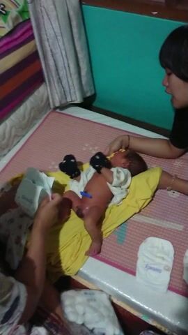 The video shows an indoor scene focused on a woman and a newborn baby. A woman with dark hair, wearing a black shirt and dark shorts, leans over the baby from the left side of the frame, her hands near the baby's upper body. The baby, dressed in a yellow outfit, lies on a yellow surface, possibly a changing mat or soft blanket, which is on top of a light-colored mat or table. A blue clip is visible on the baby's umbilical cord, indicating it is a very young infant.

On the right side of the frame, part of another person's arm and hand are visible near the baby's feet. Next to the baby on the light-colored surface, a white diaper and several small containers or bottles are present. The floor to the left is covered by a green mat, leading to a wooden door or frame with glass panels reflecting what appears to be a car wheel and other objects. On the far right, a patterned fabric or blanket is visible on the floor. The scene suggests a moment of gentle interaction or care for the newborn in a home environment.