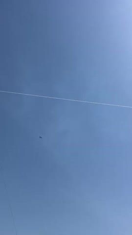 A small airplane is seen flying across a bright blue sky. A thin white line, possibly a power line or a contrail, stretches diagonally across the frame. The sun is visible in the upper right corner of the frame, casting a bright light and causing a lens flare. The airplane is dark and appears to be a propeller-driven aircraft. It is in the lower left quadrant of the frame and appears to be moving from left to right.