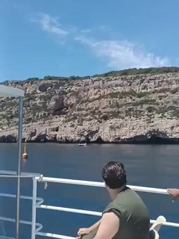 A group of people are on a boat. The boat is moving through the water, with a rocky cliff face on the left side of the frame. The sky is clear and blue. The people on the boat are looking out at the scenery. Some are standing, others are sitting. The boat has a white railing. There is a yellow buoy in the water.