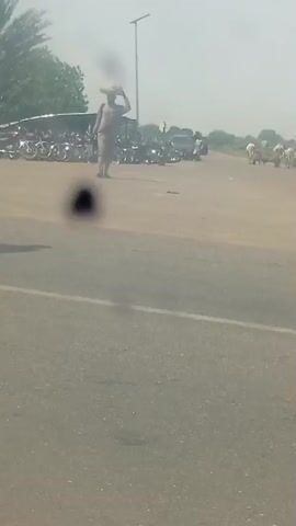 The video shows a person walking across a road carrying a bundle on their head. In the background, there are several motorcycles parked and people standing around. Further in the distance, there appear to be some cattle or other animals, also with people around them. The environment looks like an outdoor, possibly rural or semi-urban area with a road and open space. The lighting suggests it is daytime.