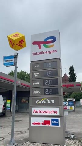 This is a vertical video taken at a gas station. A tall sign displaying fuel prices and services dominates the center of the frame. The sign has the "TotalEnergies" logo at the top. Below it, rows of digital displays show prices for Diesel (1.909), Super E10 (1.979), Super (2.039), and Super Plus (2.209). Further down, there are advertisements for "excellium," "Autowäsche" (car wash), and "AdBlue," with accompanying graphics of a car and a truck.

To the left of the main sign, a pole holds a yellow cube-shaped sign with the "Lotto" logo, and below that, a blue sign with "Hermes PaketShop" (Hermes Parcel Shop). The gas station canopy is visible in the background, along with some parked cars and trees. The ground is paved with asphalt and gravel. The sky is overcast. The video appears to be a stationary shot, with no discernible movement of people or objects within the frame.