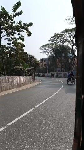 The video shows a street scene in what appears to be a South Asian country. The view is from a moving vehicle, possibly a rickshaw or auto-rickshaw. The road curves gently. Along the left side of the road is a low fence constructed from vertical wooden or metal planks, behind which is dense vegetation. Several people are walking on the road, including two men who cross the street at a diagonal.  Various vehicles, primarily rickshaws, are visible in the distance, traveling in both directions. The sky is overcast and the light is diffused. Trees are plentiful on both sides of the road. The focus is on the road and the traffic along it.