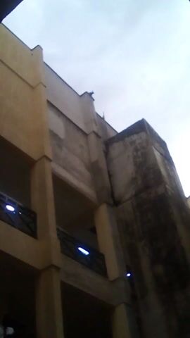The image captures an upward view of a building against a cloudy sky. The building, which appears to be multi-storied, features a combination of light yellow and gray concrete. Balconies with dark wooden railings are visible. One side of the building has a darker, weathered appearance, possibly due to age or exposure to the elements. The overall composition gives a sense of architectural detail and height.