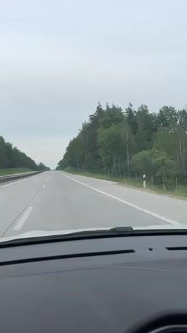 A POV shot from inside a moving car shows a multilane highway with a forest on the right, under a cloudy sky.