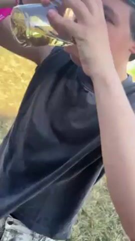 Here is a description of the video:

The video shows a person at an outdoor event, possibly a festival. The person, wearing a dark sleeveless shirt, camouflage pants, and a gray cap, is shown drinking from a clear glass. The liquid inside the glass appears to be a light amber color. In the background, there are several portable toilets (porta-potties), and other people are seen milling around. The setting is on a grassy field under a bright, sunny sky. The person is holding the glass with both hands, tilting it to drink while partially covering their face with their hands. The camera angle shifts to show the pants with a small white object in the pocket. The person also has a pink wristband on their left wrist.
