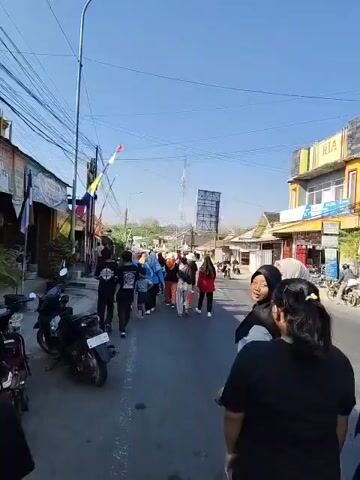 A group of people are walking down a paved road lined with buildings and parked motorcycles. The sky is clear and blue. The people are walking in the same direction, and most of them are wearing dark clothing. Some of the people are wearing headscarves. One person in the foreground is wearing a black t-shirt and has their back to the camera. Another person in the foreground is wearing a black t-shirt and a blue headscarf. There are utility poles and wires visible in the background.
