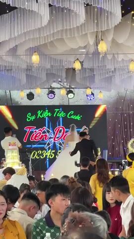 An indoor wedding reception is underway at a venue in Bình An, Vietnam. The scene features the bridal couple on a brightly lit stage. The bride, in a white wedding gown, and the groom, in a black suit, are standing together, with the groom holding a bouquet. To their right, a female emcee or singer in a light yellow dress is speaking into a microphone. Behind them, a large LED screen displays the Vietnamese text "Sự Kiện Tiệc Cưới" (Wedding Event) and "Tiến Danh," alongside a partially visible phone number "03345." A multi-tiered wedding cake is visible to the left of the stage. The ceiling is adorned with draped white fabric and numerous hanging decorative lights. Below the stage, a crowd of guests is seated at tables, observing the proceedings, contributing to a festive and joyful atmosphere.