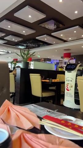 A robotic server with a cat-like face on its screen navigates a dimly lit restaurant. The robot has a white body with a tray system on the front. The robot's screen displays a pair of animated eyes and a mouth. The robot's body is adorned with the restaurant's logo, which includes Chinese characters and a red fish design. A table in the foreground is set with pink napkins, chopsticks, and a small dish of sauce. The restaurant has a modern interior with dark brown walls, a reflective ceiling with recessed lighting, and beige chairs. In the background, other tables are visible, some with patrons. The robot is moving past a table with food on its tray, suggesting it is delivering an order.