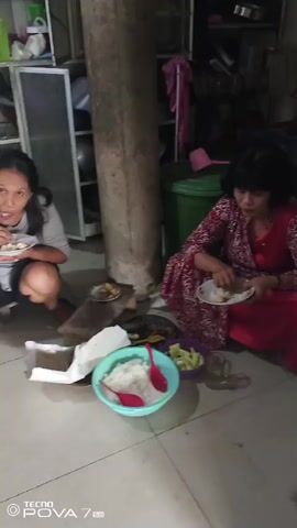 Two women are eating on the floor. One woman is wearing a red dress with a white floral pattern and is holding a plate of food. The other woman is wearing a grey shirt and is also holding a plate of food. In the background, a woman is holding a baby in a sling. She is in a kitchen with a stove and cabinets. There are also some food items on a counter.