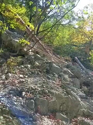 The video shows a landslide in a wooded area. Rocks and debris are scattered across the ground, and trees are leaning precariously. Sunlight filters through the leaves, casting shadows on the scene. The ground is uneven and rocky, with some patches of dirt and fallen leaves. The landslide appears to have occurred on a slope, and there is evidence of disturbed earth and vegetation. The area seems to be a natural environment, possibly a park or a hiking trail.