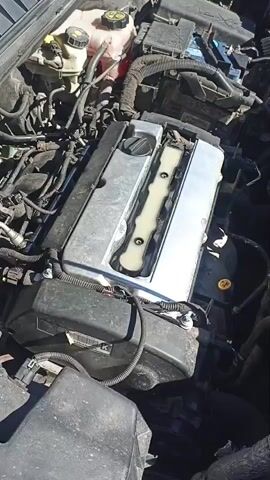 This is a top-down video of a car engine. The engine appears to be a four-cylinder engine, with a silver-colored valve cover. There are various hoses and wires connected to the engine. The engine is located in the engine bay of a car. The video is shot in daylight, and the environment appears to be an outdoor setting. The engine is not running in the video.