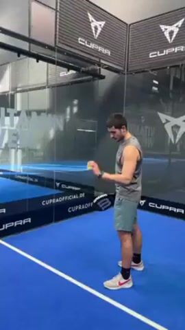 A man is playing padel tennis in an indoor court. He is wearing a gray tank top and light-green shorts with red and white sneakers. He tosses the ball up with his left hand and then swings a black and white padel racket to hit the ball. The court has blue flooring and glass walls with black banners with white “CUPRA” logos. There are also white inscriptions on the glass wall that say “VITAMIN WELL”. The video is shot at eye level.