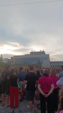 A vertical video captures a crowd of people gathered outdoors, likely for an event or festival. The subjects include a diverse group of men, women, and children, many of whom appear to be dressed in traditional folk costumes, particularly the women in long, patterned dresses and headwear. Some individuals are wearing modern casual clothing.

The scene is set in an urban or semi-urban environment, with several multi-story buildings visible in the background, suggesting a town or city setting. One prominent building in the middle ground is a modern structure with a glass facade and antennae on its roof. In the distance, more buildings and trees can be seen. The ground appears to be paved, with some industrial-looking elements like a large metal ring and a thick rope visible in the foreground.

The time of day seems to be late afternoon or early evening, with the sun setting or low in the sky, casting a soft light and creating a warm, possibly slightly hazy atmosphere. The sky is partly cloudy, with streaks of light breaking through. There are no obvious vehicles directly in the crowd's vicinity, though some red vehicles, possibly fire trucks or emergency vehicles, are visible further back.

The activity taking place is a gathering or celebration. The presence of traditional attire suggests a cultural event, possibly a folk festival or local celebration. The mood of the crowd appears to be festive and engaged, with people standing and observing. There are no clear indications of any organized performance or specific activity happening at the exact moment the video was recorded, but the setup suggests anticipation.

There is no visible text in the video. The location context provided is Криводановка, Russia, which aligns with the visual cues of the architecture and the possibility of a traditional Russian folk event.