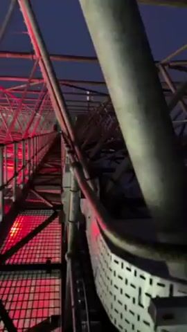 The video shows the exterior of a metal structure illuminated with red lighting. The sky is dark and some city lights are visible in the distance. The structure has a crisscross design made up of various tubes and bars. The camera appears to be moving up the structure, showing a low angle view, and later shifts to a slightly higher angle showing more of the cityscape.